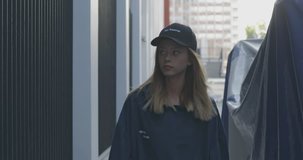 Trendy girl wearing cap and blue sweatshirt goes down alley and looks into camera.. City urban style. 4K video shooting by handheld gimbal