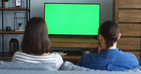 Back view on the young couple sitting in the living room and watching TV with green screen, men changing channels with a remote control. Chroma key. Indoors