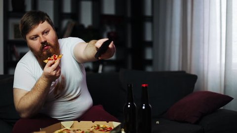 Weak-willed overweight man eating pizza and drinking beer sitting on the couch. Concept of malnutrition, sport, obesity