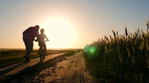 Dad is teaching daughter how to ride bicycle on the meadow at sunset time. Happy father rejoices that her daughter learned to ride a bike.