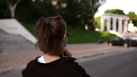 Backside view of a young modern girl in black casual walking by park or street. Looking back over her shoulder and smiling. Puts on wareless headphones. Leisure time