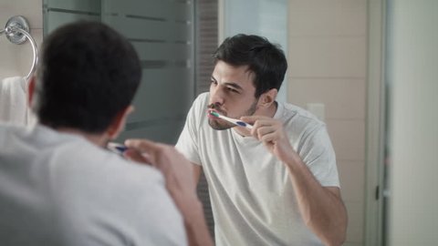 Hispanic person with beard grooming in bathroom at home for morning routine and body care. Adult metrosexual man brushing teeth and looking at mirror