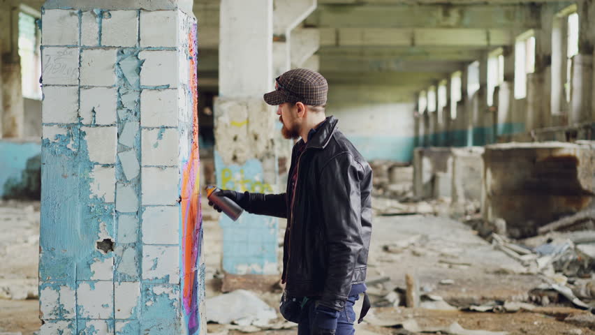Side view of graffiti artist bearded guy drawing on damaged column inside emply industrial building using bright aerosol paint. Creativity and people concept.