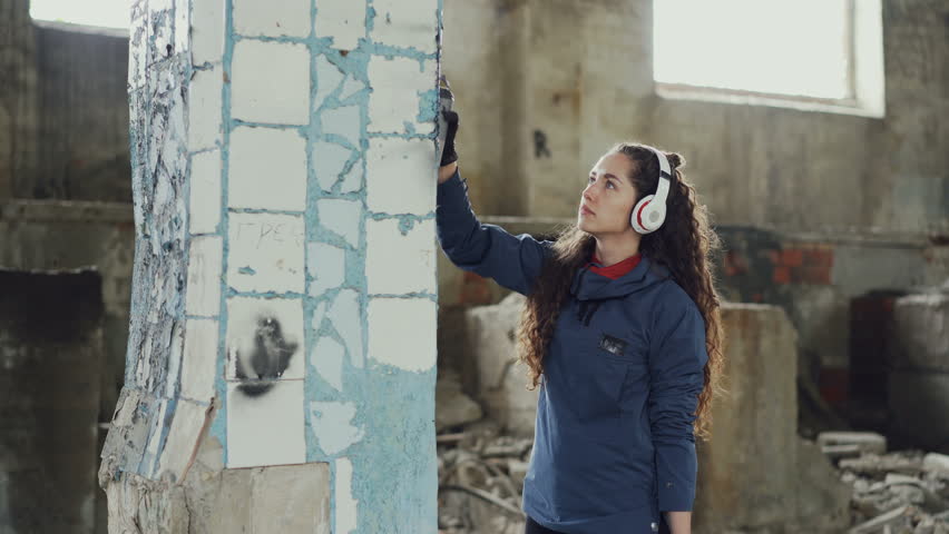 Attractive girl is concentrated on decorating old dirty column with graffiti in abandoned building using aerosol paint. Young woman is listening to music with headphones.