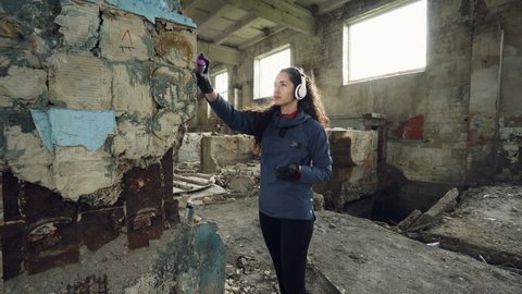 Pretty young woman graffiti artist is painting with spray paint inside old warehouse and listening to music through wireless headphones. Youth subculture concept.