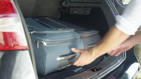 SLOW MOTION, CLOSE UP: Unknown male lifting heavy travel bags out of the spacious trunk of his big black car. Unrecognizable young man unpacking luggage out of his SUV after awesome summer vacation.