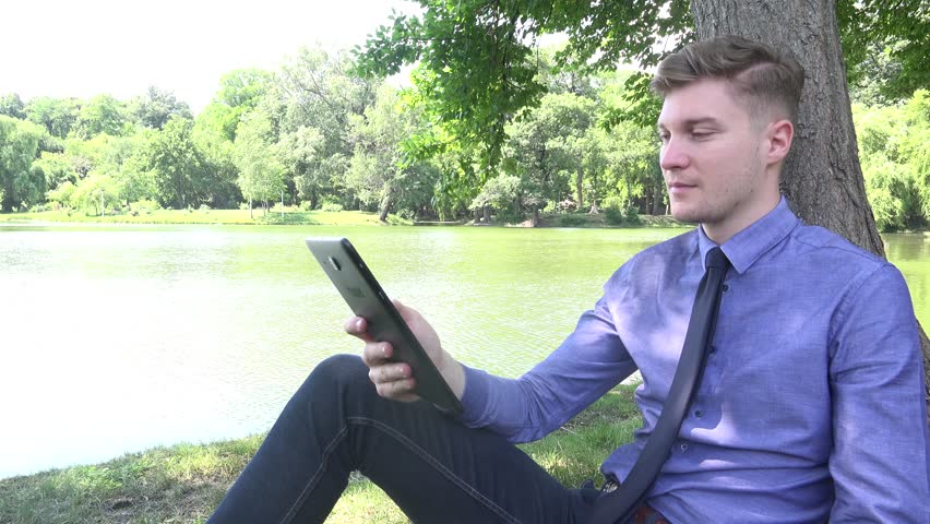 Portrait young businessman sitting by lake tree watching tablet computer smiling