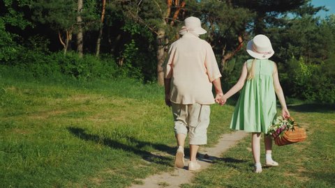 Grandmother is walking with her granddaughter in the forest. Holding hands, the girl carries a basket with wild flowers. Active seniors, rear view video