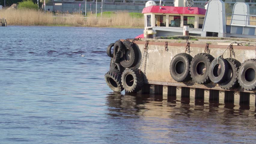 Line of black tires hanging on the side of the port with the sailboats and cargo ships docking on the side