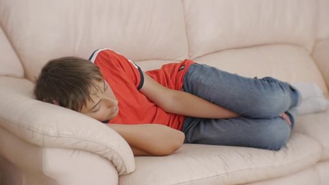 Teenager sleeping on couch. Caucasian teen boy in red t-shirt and blue jeans sleeping on beige leather sofa in daytime. Lack of sleep