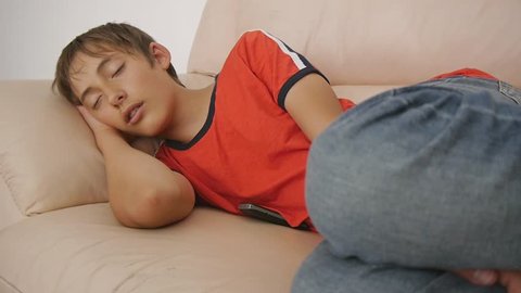 Teenager sleeping on couch. Caucasian teen boy in red t-shirt and blue jeans sleeping on beige leather sofa in daytime. Lack of sleep