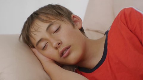 Teenager sleeping on couch. Close-up of caucasian teen boy in red t-shirt sleeping on beige leather sofa in daytime. Lack of sleep