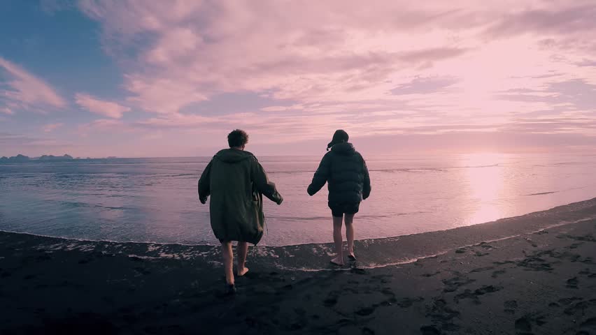Two best friends or couple in winter jackets run on beach in cold shallow waters,jump and laugh, make silly faces and movements,happy to spend time together in vacation on pink sunset,active lifestyle