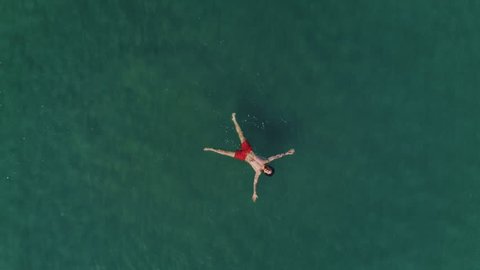 Aerial drone birdseye view shot of man in bright red swimming shorts lay flat on water surface of ocean sea, crystal clear see through turquoise waves, relaxed and chill vacation holiday deserved time