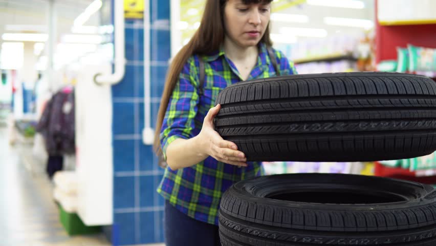 A young woman in a blue checkered shirt chooses and buys car tires at the supermarket in the department of automotive goods.
