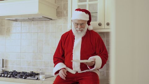 Santa sits on kitchen furniture and eats cookies with milk
