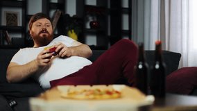 Fat man plays the game console. He is sitting on the couch in his apartment drinks beer and eats fast food. Concept of video games and malnutrition, obesity