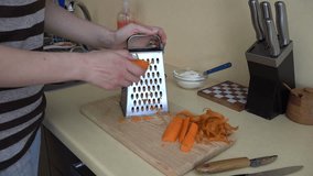 Housewife woman hand grate peeled orange carrot with shredder tool on wooden cutting board in kitchen. Static closeup shot. 4K UHD video clip.