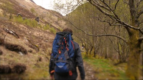 Young traveller hiking through the Scottish Highlands. Man path walking and back packing trekking through natural landscapes of mountains and trees with bag. British hiker travelling in Scotland.
