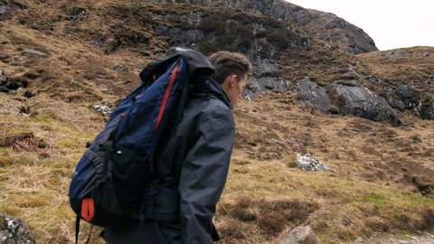 4K Young traveller hiking alone through the Scottish Highlands. Man on path walking and back pack trekking through rural mountains. A natural rugged landscape with a British tourist location. 