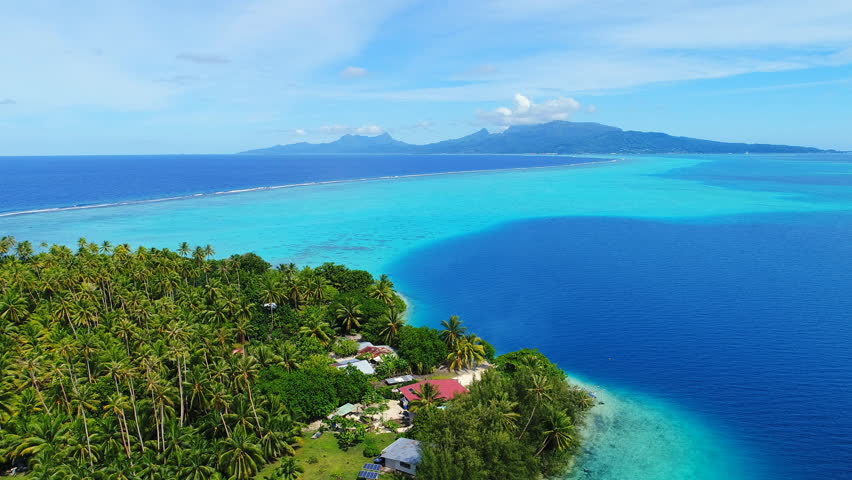 Aerial view of coral barrier reef of Tahaa island, palm trees on islet Motu Atger and clear water of blue lagoon, Raiatea on background - South Pacific Ocean, seascape of French Polynesia from above