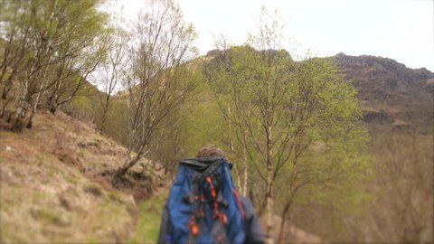 4K Young traveller hiking through the Scottish Highlands. Man path walking and back packing trekking through hilly natural landscapes of mountains and trees with bag. British hiker travelling