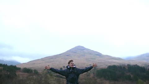 4K Young traveller dancing and looking at the view of a mountain in the Scottish Highlands. An ecstatic student back packing and nature trekking through rural landscape. A British tourist exploring 