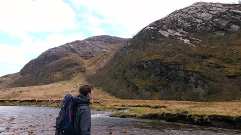4K Young traveller in Scottish Highlands. Student back pack nature path trekking through mountains and lakes. A natural marsh and water landscape with a British tourist exploring rural Scotland.