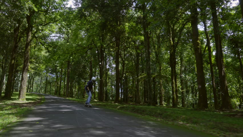 Front view follow shot of active young man in helmet riding skateboard in green park on warm summer day