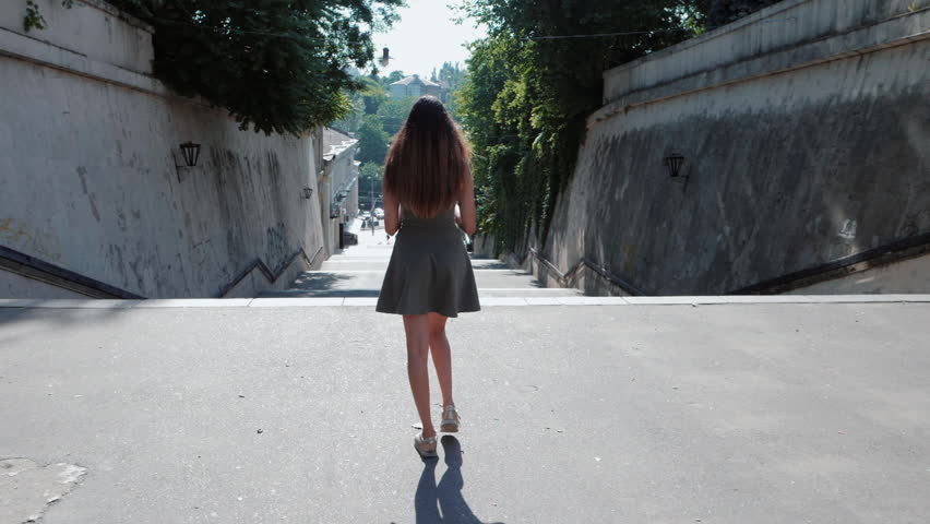 Young attractive girl in short dress and sneakers goes down big stairs of city. Woman with ombre dyed long hairstyle. Concept of travelling, tourism. Back view. Slow motion.