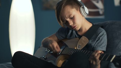 Boy sitting on bed at home and listening to music in headphones while practicing guitar at night