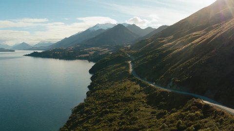 Road trip in New Zealand countryside beside blue lake. White van/campervan driving down long winding road.