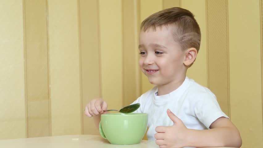 Kid with pleasure and with a smile eating soup from a plate with a spoon, sitting at the table.
