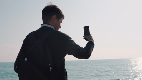 Traveler is filming calm seascape in sunny day, standing back to camera. He is holding mobile phone in hands and moving it, solar path on a sea surface