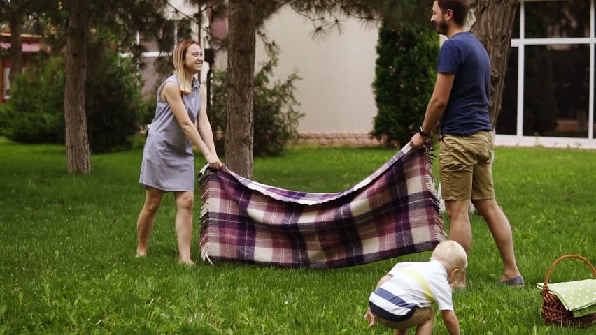 Slow motion of a young family with a son on a green lawn. Parents are spreading the plaid. Excitement. Blonde little boy on a forward perspective