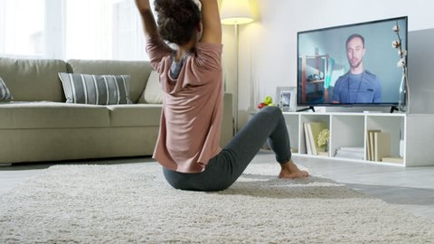PAN with slowmo  of young woman exercising at home with personal fitness trainer by video call: she looking at TV screen and doing crunches with raised arms on floor