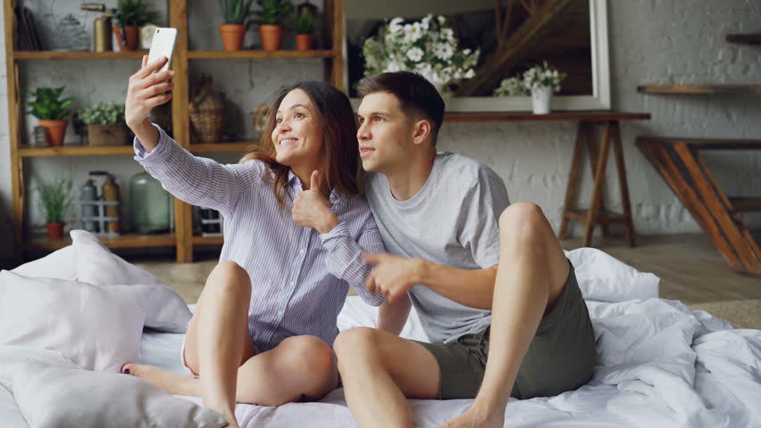 Modern married couple is taking selfie in bedroom gesturing posing and kissing while sitting on bed together. Modern technology and people concept.