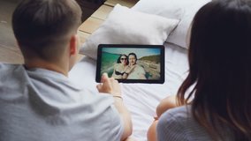 Married couple is talking to friends on tablet, happy lovers are laughing and kissing on screen with beautiful sea in background. Video calls and communication concept.