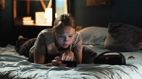 Young casual woman lying on bed alone and watching social network on smartphone relaxing alone at night