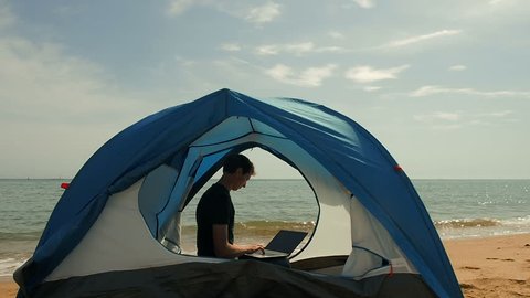 Freelancer Man Working Using Laptop Sitting In A Camping Tent On The Beach. Freelancer working on new startup project using laptop computer and wireless connection. Freelance Summer Travel 