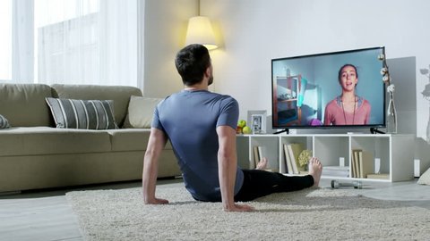 PAN of bearded man in sportswear doing knee tuck crunches on floor of his apartment and looking at TV screen as online personal fitness trainer instructing him by video call