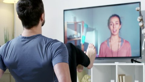 PAN of man in sportswear doing knee tuck crunches and looking at TV screen with female online personal trainer giving him instructions and counting