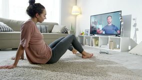 PAN of young woman in sportswear doing push-ups on floor on her apartment as male online personal trainer counting time on stopwatch and giving instructions by video call on TV screen