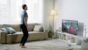 Wide shot of laughing bearded man in sportswear doing scissor jumps and jumping jacks while looking at cheerful online person coach on TV screen