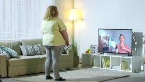 Medium shot of overweight woman in sportswear looking at online personal trainer on TV screen and doing marching exercise in living room