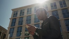 Medium shot of young African American man using his smartphone while waiting for somebody near building in sunlight and looking in distance