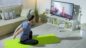 High angle shot of bearded man sitting in lotus pose on yoga mat and stretching arms and sides while following instructions of female online personal trainer on TV screen