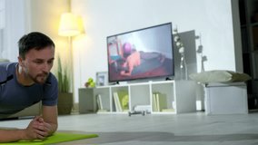 Medium shot bearded man in sportswear doing plank on mat, then resting and breathing hard while training with online personal coach by video call