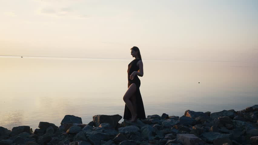 Carefree woman in long black dress dancing on beach at sunset.