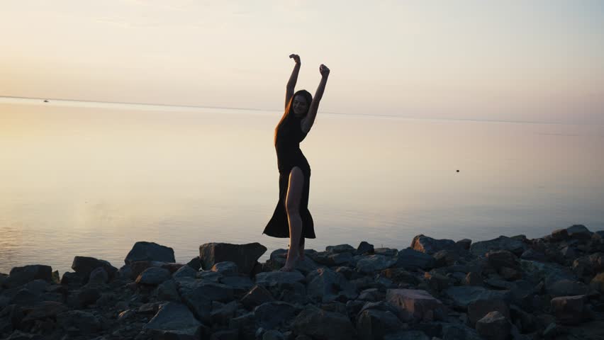 Carefree woman in long black dress dancing on beach at sunset.
